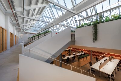 Zona de trabalho em open space com mesas de madeira clara, divisórias baixas e cadeiras ergonômicas. A luz natural entra através de grandes janelas, criando um ambiente moderno e minimalista, com toques de design japonês.