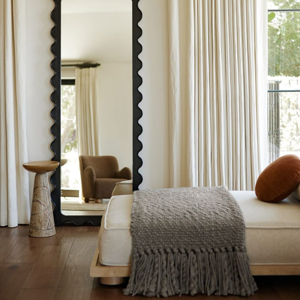 Interior minimalista com espelho de moldura ondulada e banco estofado em tons neutros, criado através de um design de interiores pensado para promover bem estar e reforçar a saúde e bem estar. A luz natural e as texturas suaves contribuem para a saúde mental e intensificam o bem estar, mostrando como o design de interiores pode apoiar a saúde e bem estar e favorecer um ambiente que nutre a saúde mental.