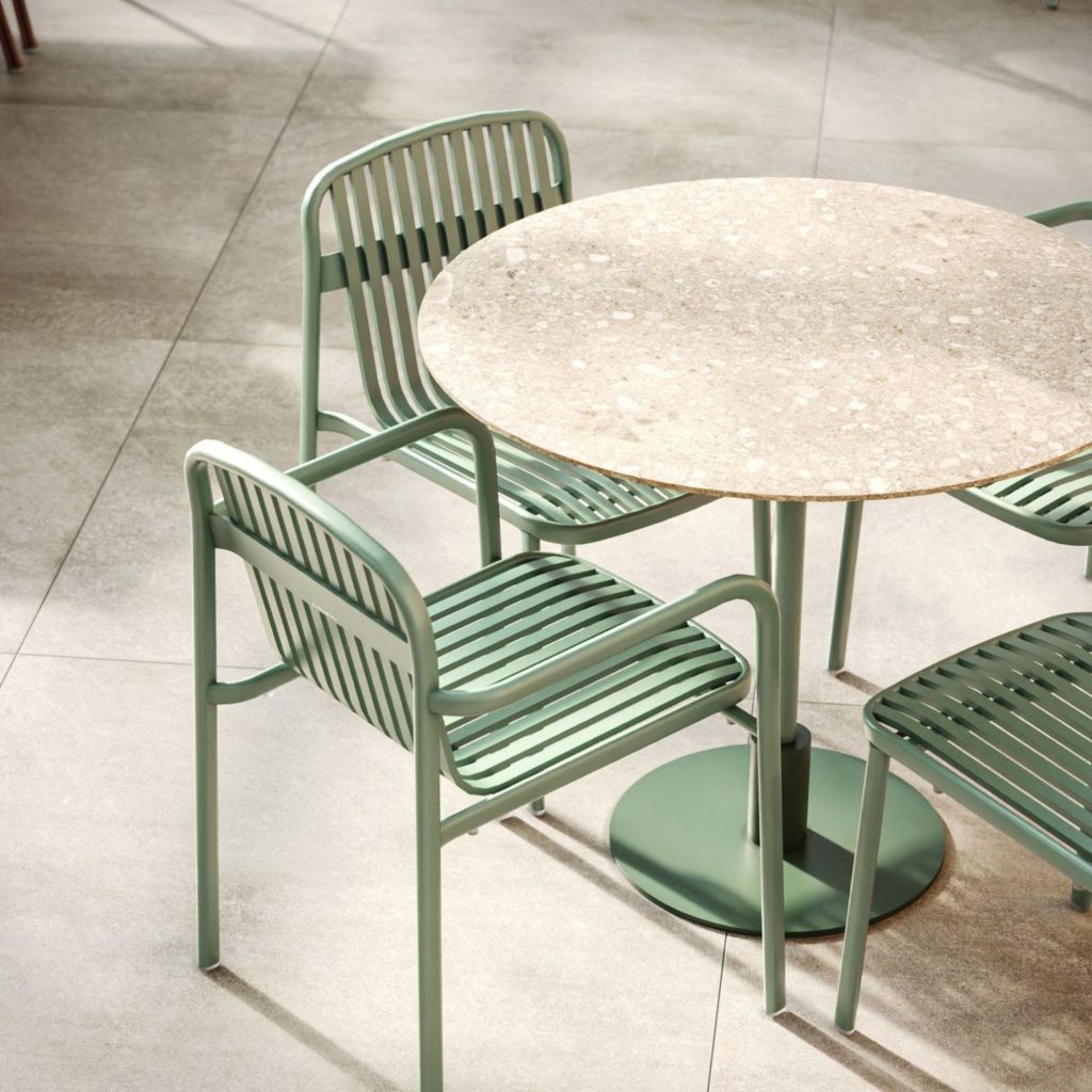 Conjunto de mesa redonda com tampo em terrazzo e cadeiras metálicas verdes, inserido num ambiente contemporâneo onde detalhes em cor pastel reforçam a leveza do design de interiores. As linhas simples e o verde suave das cadeiras aproximam-se de uma paleta de cores pastel, criando um cenário funcional e elegante. O design de interiores aqui presente demonstra como a cor pastel pode ser aplicada de forma subtil, enquanto a paleta de cores pastel contribui para um ambiente visualmente coeso e moderno.
