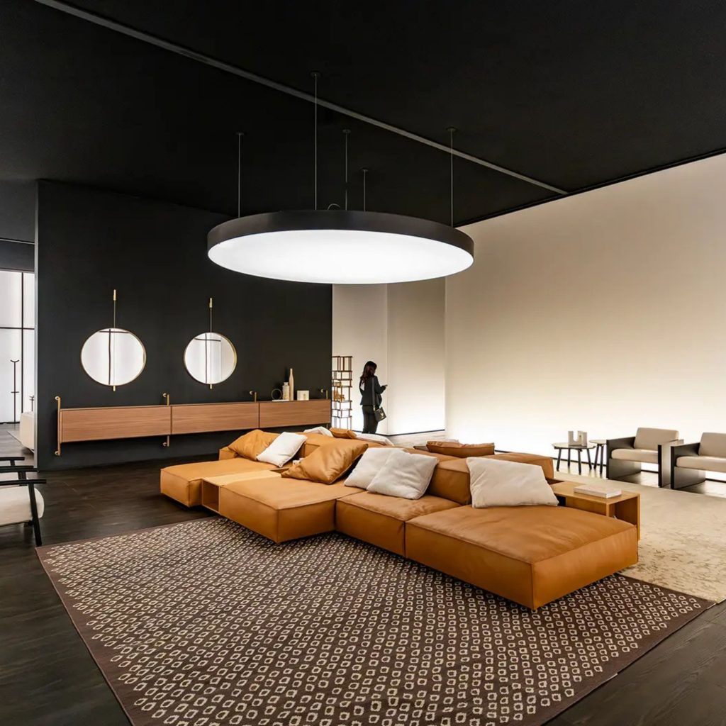 Sala moderna com sofá caramelo e iluminação circular difusa, inspirada no ambiente do salone del mobile 2026 e nas suas tendências. O contraste entre paredes claras e escuras demonstra como o salone del mobile 2026 molda o design de interiores, antecipando tendências que combinam conforto visual e linhas contemporâneas no design de interiores.