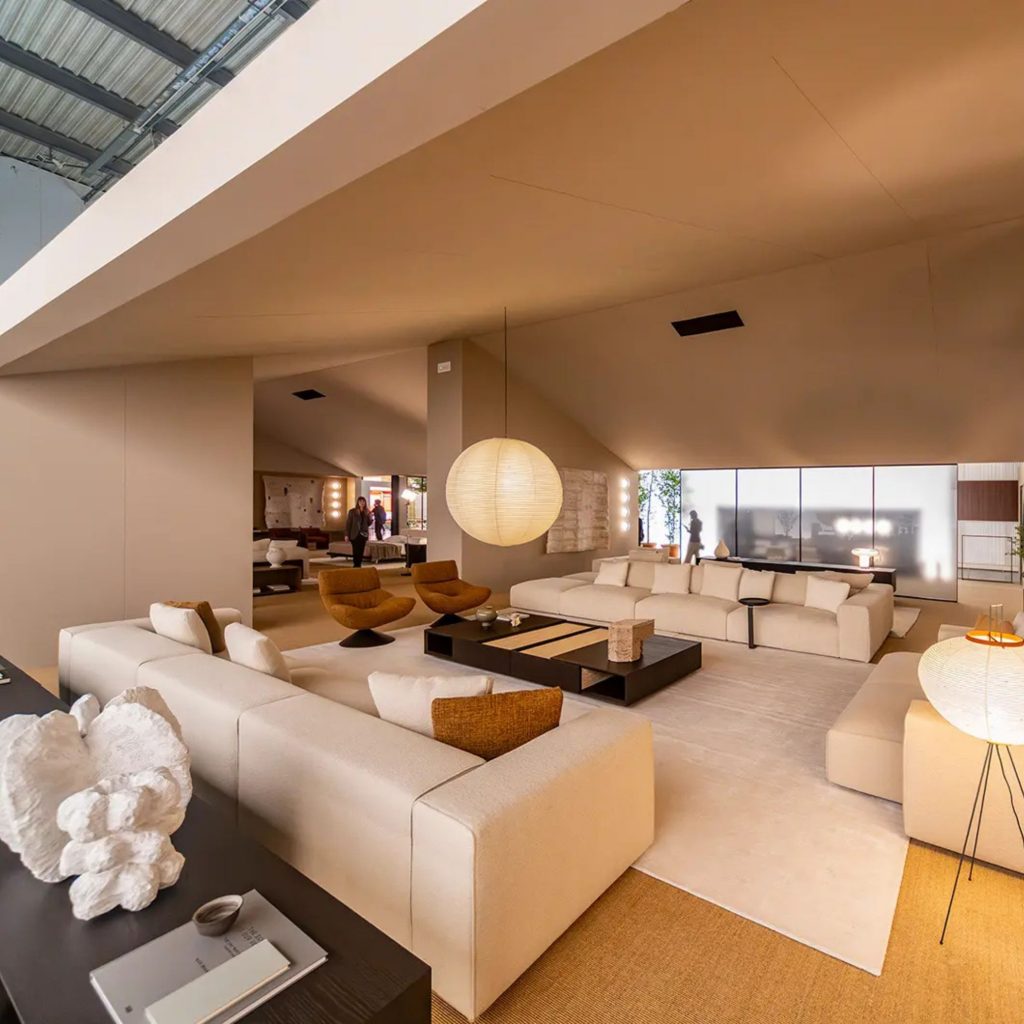 Sala ampla com sofás modulares, cadeirões e iluminação suave, refletindo influências do salone del mobile 2026 e das suas tendências. O ambiente contemporâneo mostra como o salone del mobile 2026 continua a orientar o design de interiores, destacando tendências que privilegiam conforto, proporção e harmonia no design de interiores.