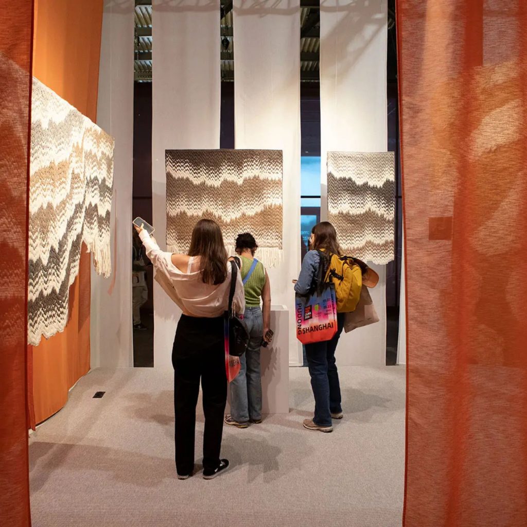 Exposição têxtil com painéis de tecidos artesanais e visitantes a observar detalhes, evocando o espírito do salone del mobile 2026 e das suas tendências. A paleta quente e as texturas revelam como o salone del mobile 2026 influencia o design de interiores, reforçando tendências que valorizam materiais naturais e expressão manual no design de interiores.
