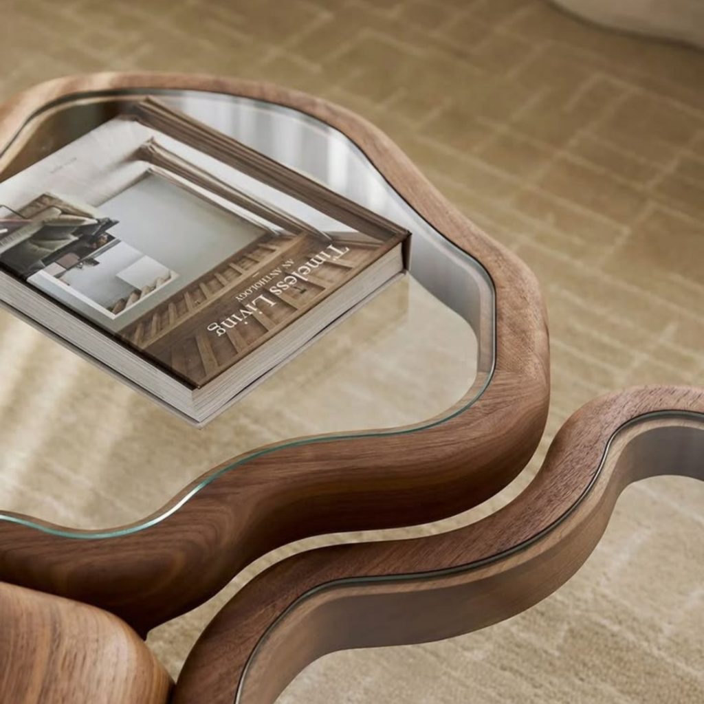A mesa de centro em madeira curva e tampo de vidro destaca-se numa decoração que privilegia formas orgânicas e materiais naturais, criando um ambiente que desperta emoções de calma e contemplação. A presença do livro reforça a ligação entre espaço e narrativa, revelando como a arquitetura influencia a forma como nos relacionamos com os objetos. A suavidade da composição traduz uma decoração sensorial, onde as emoções são moldadas pela intenção espacial e pela coerência da arquitetura, que aqui se manifesta em linhas fluidas e proporções equilibradas.