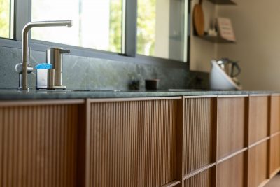 Cozinha moderna com bancada escura e arrumação ripada que reforçam a estética depurada da cozinha moderna; A luz natural valoriza a composição e acentua a presença de uma cozinha moderna com ilha; Ambiente funcional onde cada detalhe contribui para a leitura contemporânea da cozinha moderna e da cozinha moderna com ilha.