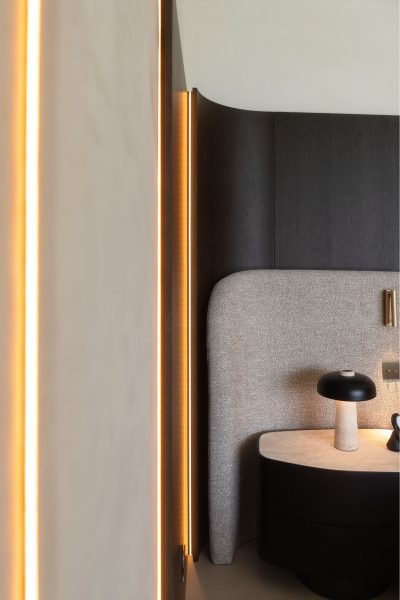 Decoração de quarto com iluminação quente que envolve a zona da cama de casal. A mesa de cabeceira em madeira escura reforça a elegância da decoração de quarto. Composição minimalista onde a textura suave da cabeceira valoriza a cama de casal, acentuando a coerência da decoração de quarto.
