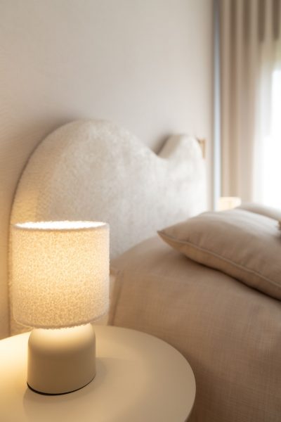Candeeiro de mesa junto a cabeceira estofada em tecido bouclé num quarto de criança. A decoração destaca texturas suaves, a decoração cria uma atmosfera confortável e a decoração contribui para um ambiente acolhedor. O design de interiores explora materiais táteis, o design de interiores privilegia a luz indireta e o design de interiores reforça o bem-estar. Este quarto de criança foi concebido para momentos de descanso, tornando o quarto de criança mais confortável, o quarto de criança mais sereno e o quarto de criança mais convidativo.