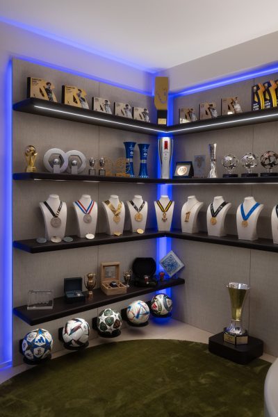 Estantes iluminadas com medalhas, troféus e bolas autografadas integradas numa área de exposição personalizada. A decoração organiza os elementos de forma harmoniosa, a decoração valoriza cada conquista e a decoração reforça o carácter exclusivo do espaço. A personalização orienta toda a composição, a personalização transforma memórias em peças centrais e a personalização confere autenticidade ao ambiente. Este projeto de design de interiores privilegia a apresentação das coleções, o projeto de design de interiores recorre à iluminação para destacar cada objeto e o projeto de design de interiores cria uma experiência visual envolvente. Integrado num projeto de design de interiores de luxo, este ambiente demonstra como um projeto de design de interiores de luxo pode unir sofisticação e significado pessoal, sendo um marcante projeto de design de interiores de luxo.