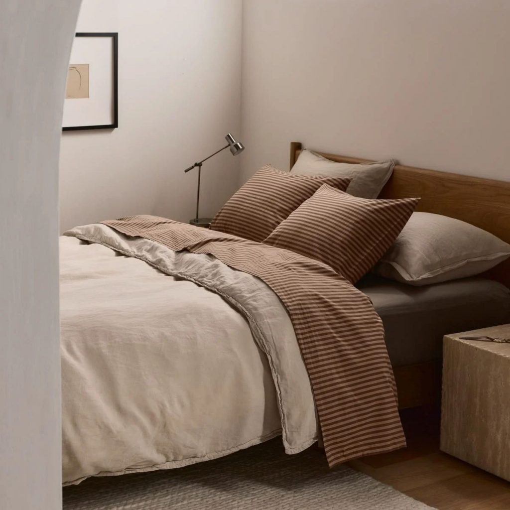 Cama de casal vestida com lençóis claros e lençóis flanela às riscas castanhas, combinados com almofadas e cabeceira em madeira.