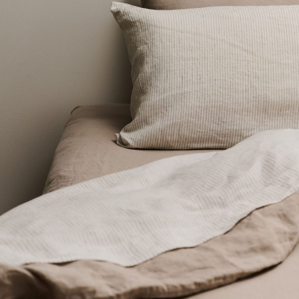 Cama de casal com lençóis em tons neutros, detalhe de almofada riscada e sobreposição de lençóis flanela em bege.