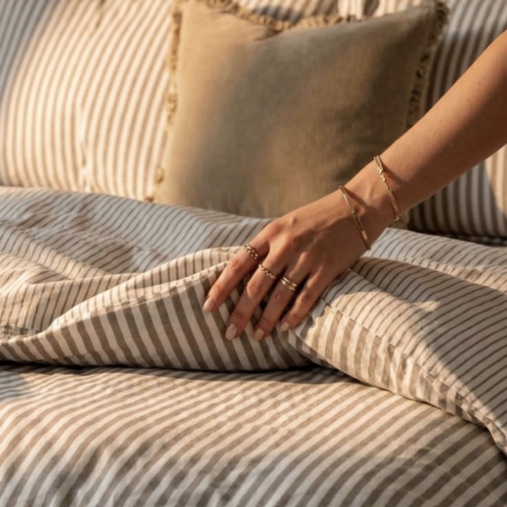 Mão a tocar no tecido suave de uma capa de edredão às riscas que veste um confortável edredão sobreposto por uma colcha leve como inspiração de colcha para cama de casal