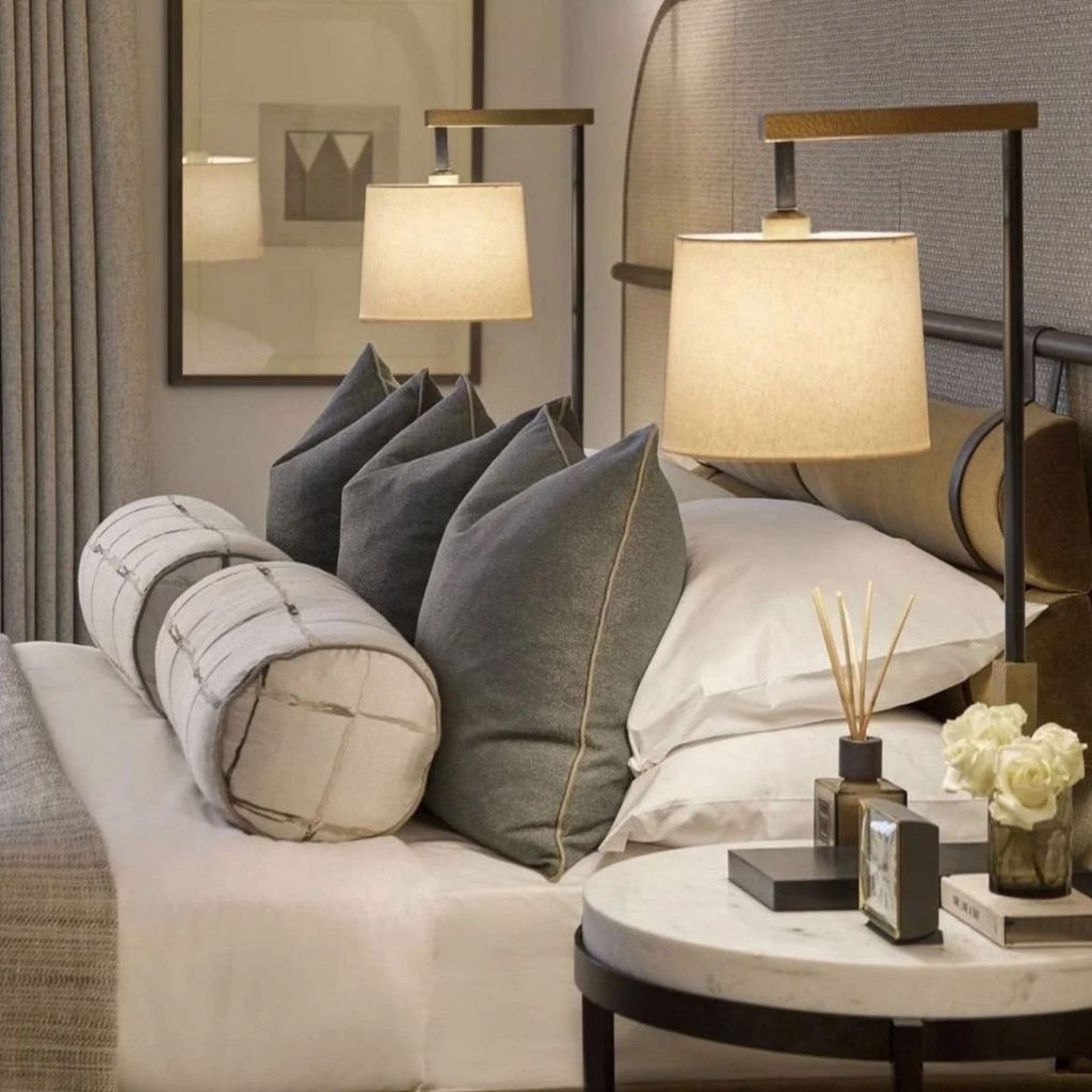 Arrumação cuidada de almofadas decorativas para cama em tons cinzentos e rolos estampados sobrepostos a almofadas brancas num quarto elegante com almofadas decorativas de veludo e tecido texturado.