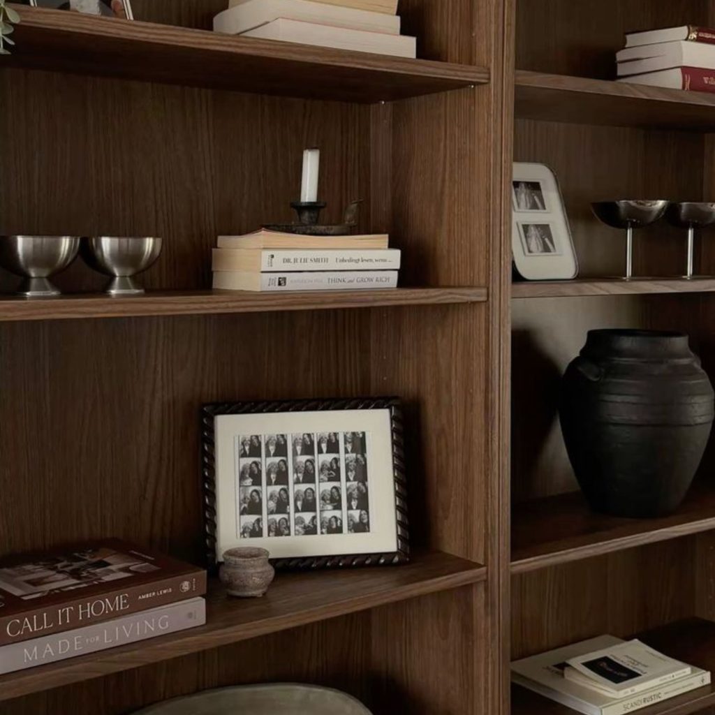 Pormenor de decoração e design de interiores numa biblioteca em casa, exibindo uma elegante estante de madeira com várias estantes para livros, vasos e molduras decorativas junto a um recanto com cadeirão.
